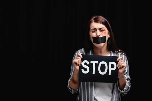 Offended woman with adhesive tape on mouth and bruises holding card with stop lettering isolated on black