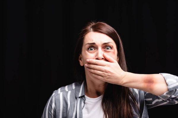 Frightened woman with bruises covering mouth with hand isolated on black 