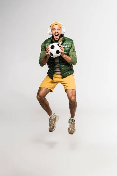 Excited Sportsman Headphones Jumping Holding Football Grey Background — Stock Photo, Image