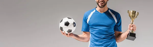 灰色のバナーに隔離されたチャンピオンカップとサッカーを保持する笑顔のスポーツマンの作物ビュー — ストック写真