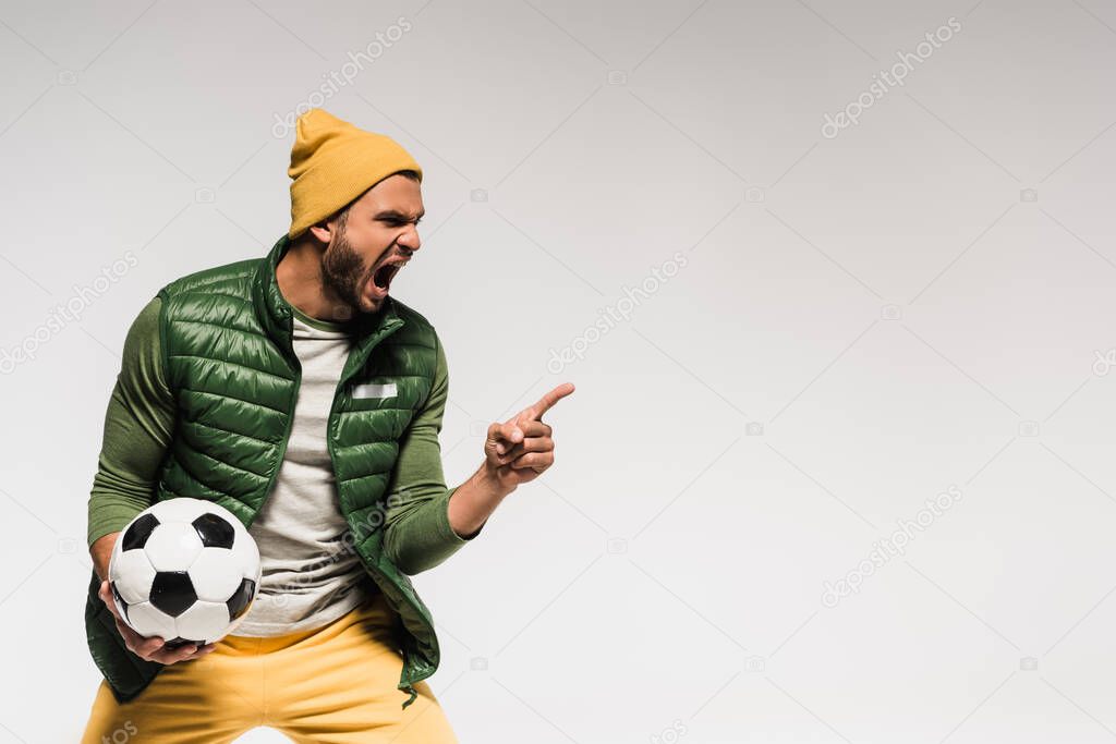Aggressive sportsman holding football while screaming and pointing with finger isolated on grey 