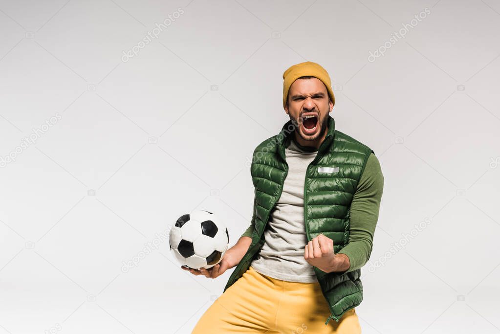 Excited sportsman with open mouth showing yes gesture and holding football isolated on grey 