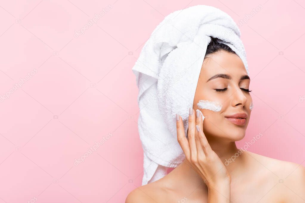 beautiful woman with towel on hair, closed eyes and cosmetic cream on face isolated on pink