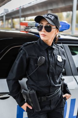 Elleri ceplerinde genç bir kadın polis, şehir sokağındaki bulanık arka planda devriye arabasının yanından uzağa bakıyor.