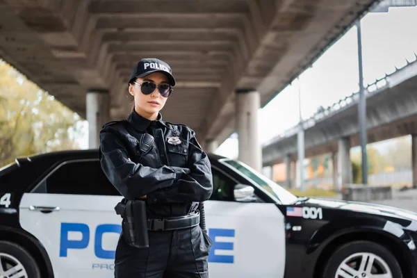 自信的女警察 双手交叉 凝视着警车旁边的摄像头 背景模糊 在城市街道上 — 图库照片