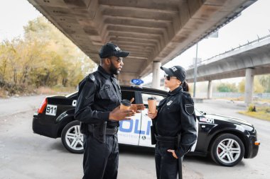 Kağıt bardaklı çok kültürlü polis memurları şehir sokaklarının bulanık arka planında devriye arabasının yanında konuşuyorlar.