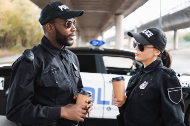 Kağıt bardaklı çok kültürlü polis memurları arka planda bulanık devriye arabasıyla konuşuyor.