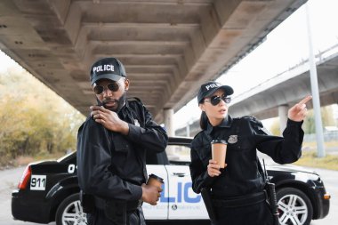 Ciddi kadın polis parmaklarıyla Afro-Amerikan polisini işaret ediyor radyoda açık alanda bulanık arka planda konuşuyor.