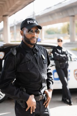 Kendine güvenen Afro-Amerikan polisi şehir sokaklarındaki bulanık arka planda kameraya bakıyor.