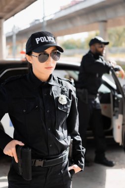 Elinde silah olan kendine güveni tam bir polis memuru arka planda bulanık Afrikalı Amerikalı meslektaşıyla yan yana bakıyor.