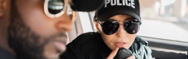 Radyoda konuşan ciddi bir polis memuru. Ön planda Afro-Amerikan polisi var. Devriye arabasında.