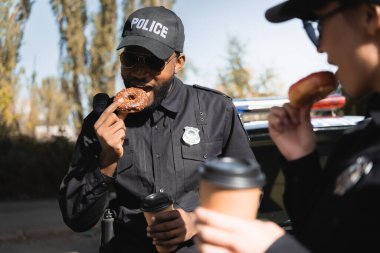 african american policeman with paper cup eating doughnut with blurred colleague on foreground outdoors clipart