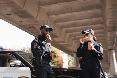 Çok kültürlü polis memurları ellerinde kağıt bardaklarla şehir sokaklarında devriye arabasının yanında donut yiyorlar.
