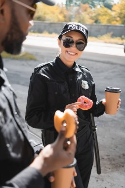 happy policewoman with doughnut and paper cup standing near african american colleague on blurred foreground outdoors clipart