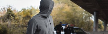 hooded offender looking away with blurred multicultural police officers on background outdoors, banner clipart