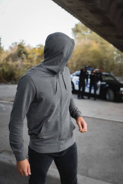 hooded thief looking away with blurred multicultural police officers on background outdoors clipart