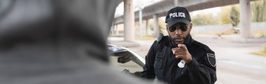 serious african american police officer pointing with finger at blurred hooded offender on foreground on urban street, banner clipart