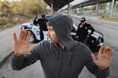 Elleri havada kapüşonlu suçlu, şehir sokaklarında arka planda bulanık, çok kültürlü polis memurlarına bakıyor.