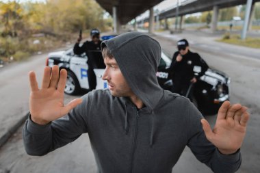 hooded offender with raised hands looking away with blurred multicultural police officers on background on urban street clipart