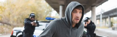 scared hooded offender running from multicultural police officers on blurred background outdoors, banner clipart