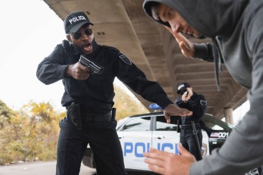 Öfkeli Afro-Amerikan polisi şehir caddesinde bulanık, teslim olmuş bir suçluyu silahla hedef alıyor.
