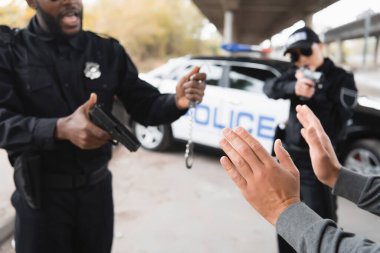 offender showing hands while multicultural police officers holding pistols and handcuffs on blurred background outdoors clipart