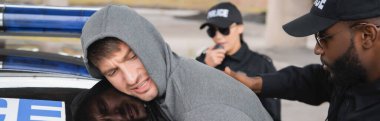 Afro-Amerikan polisi arka planda bulanık meslektaşıyla devriye arabasının yanında kapüşonlu suçluyu tutukladı.