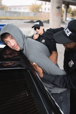 Afro-Amerikan polisi üzgün kapüşonlu suçluyu arıyor. Arka planda bulanık bir şekilde arabaya yaslanıyor.