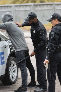 Ön planda bulanık meslektaşı ile devriye arabasının yanında kapüşonlu kapüşonlu suçluyu arayan Afro-Amerikan polisi