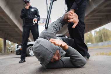Afro-Amerikan polisi kapüşonlu suçluyu tutuklayıp yüzü örtülmüş bir şekilde sokakta bulanık arka planda yatıyor.