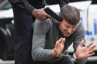 Afro-Amerikan polisi dışarıda bulanık arka planda korkmuş suçluya silah doğrultuyor.
