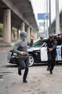 Şok olmuş Afro-Amerikan polisinden kaçan kapüşonlu bir suçlu. Arka planda bulanık bir silaha nişan alıyor.