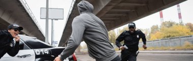 hooded offender running from multicultural police officers near patrol car on urban street, banner clipart