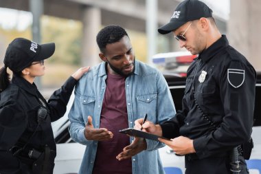 Afro-Amerikan kurbanını sakinleştiren bir polis memuru arka planda bulanık bir panoyla polisle konuşuyor.