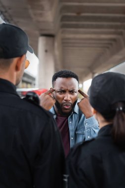 angry african american victim showing you are insane gesture while arguing with police officers on blurred foreground outdoors clipart