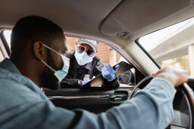 Tıbbi maskeli bir kadın, siyah bir arabanın ön camında siyah maske takan Afro-Amerikan bir sürücünün yanında pano tutuyor. 