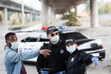 Tıbbi maskeli Afro-Amerikan kurban, lateks eldivenli ve pano takmış polis memurlarının yanında parmağıyla işaret ediyor. 