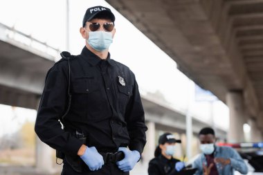 Tıbbi maskeli ve lateks eldivenli bir polis memuru dışarıda bulanık arka planda meslektaşı ve Afro-Amerikan kurbanının yanında duruyor. 
