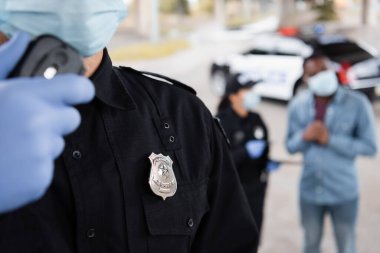 Plastik eldiven ve tıbbi maske takmış bir polis memuru, arka planda bulanık bir geçmişi olan, iş arkadaşı ve Afrikalı Amerikalı bir kurbanın yanında telsiz mesajı tutuyor. 