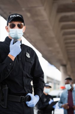 Tıbbi maskeli ve lateks eldivenli bir polis memuru meslektaşının yanında telsiz kullanıyor ve şehir sokaklarında bulanık geçmişi olan Afro-Amerikan bir adam. 