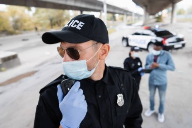 Lateks eldiven ve tıbbi maske takmış bir polis meslektaşının yanında telsiz kullanıyor ve şehir sokaklarında bulanık bir geçmişi olan Afrikalı Amerikalı bir kurban var. 