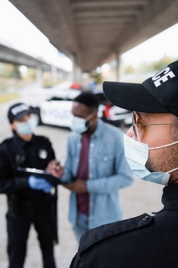 Tıbbi maskeli ve güneş gözlüklü bir polis memuru bulanık arka planda Afrika kökenli Amerikalı kurban ve meslektaşının yanında duruyor. 