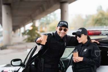 Neşeli polis memuru, iş arkadaşının elini kaldırdı. Elinde kahve, dışarıda araba var. 