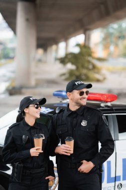 Güneş gözlüklü, gülümseyen polis memurları şehir sokaklarının bulanık arka planında otomatiğe gitmek için ellerinde kahve tutuyorlar. 