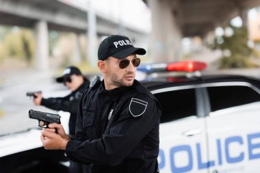 Güneş gözlüklü bir polis silah tutuyor ve bulanık arka planda meslektaşı ve arabasına bakıyor. 