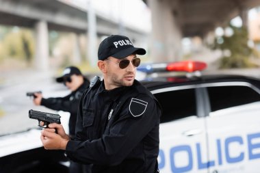 Policeman in sunglasses holding gun and looking away near colleague and car on blurred background  clipart