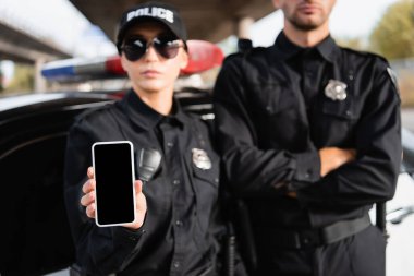 Meslektaşının yanında polis memuru ve bulanık arka planda arabası olan boş ekranlı akıllı telefon. 