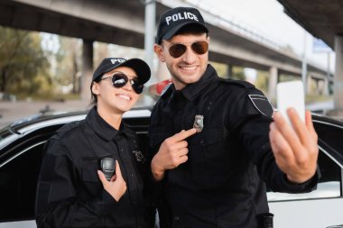 Neşeli polis memurları dışarıda bulanık ön planda akıllı telefondan selfie çekerken rozetlerini ve telsizlerini gösteriyor. 
