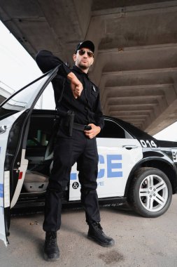 Polis memuru güneş gözlüklü ve üniformalı. Şehir sokağında arabanın yanında duruyor. 