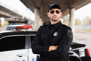 Güneş gözlüklü bir polis memuru dışarıda arka planda, arabanın yanında çapraz kollarla dikiliyor. 